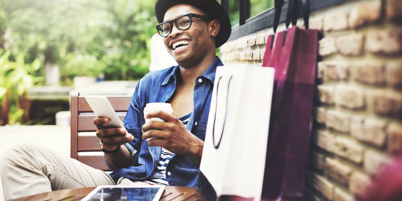 A young man sits on a patio holding a drink and his phone with shopping bags on the table